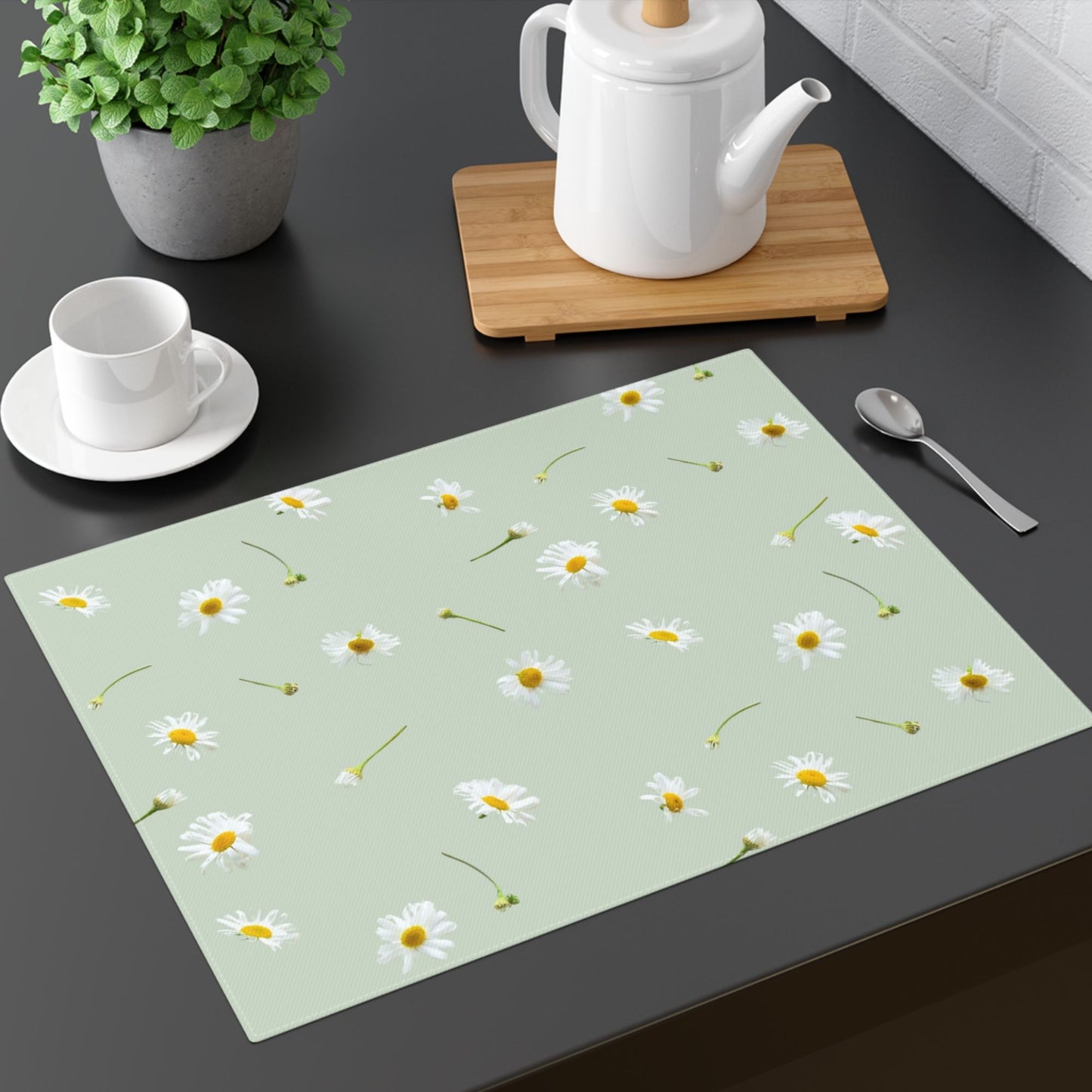  A modern black table setup with a pastel green placemat decorated with white daisies. The arrangement includes a white teapot on a wooden tray, a white teacup on a saucer.