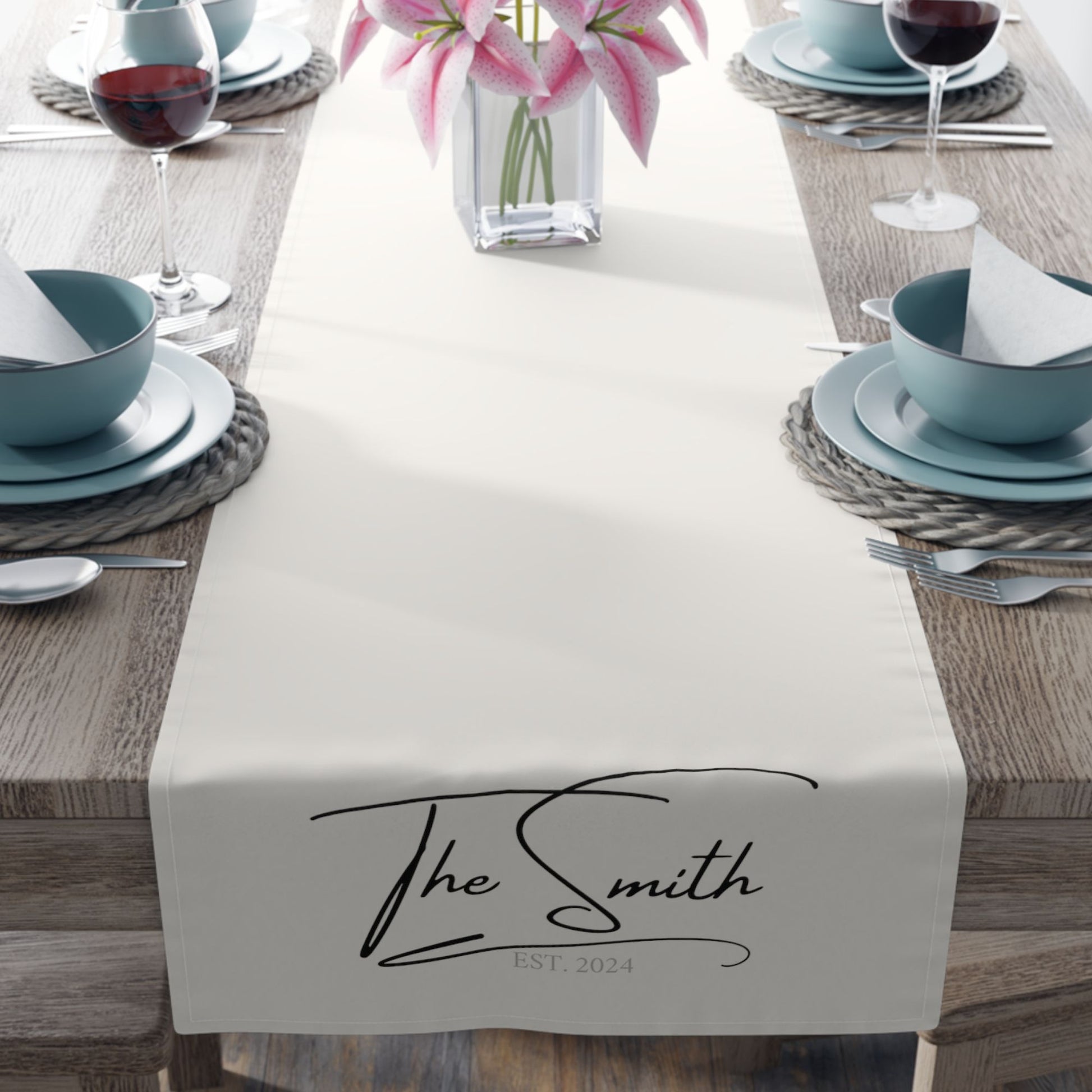 Elegant dining setup with a personalized table runner that reads "The Smith" and "EST. 2024," adorned with pink lilies and set with blue dishware.