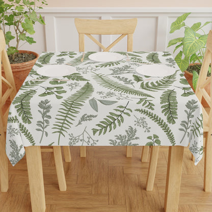 A dining table set with a white tablecloth featuring a green fern and leaf pattern, accompanied by four wooden chairs and surrounded by potted plants, creating a natural and inviting atmosphere.