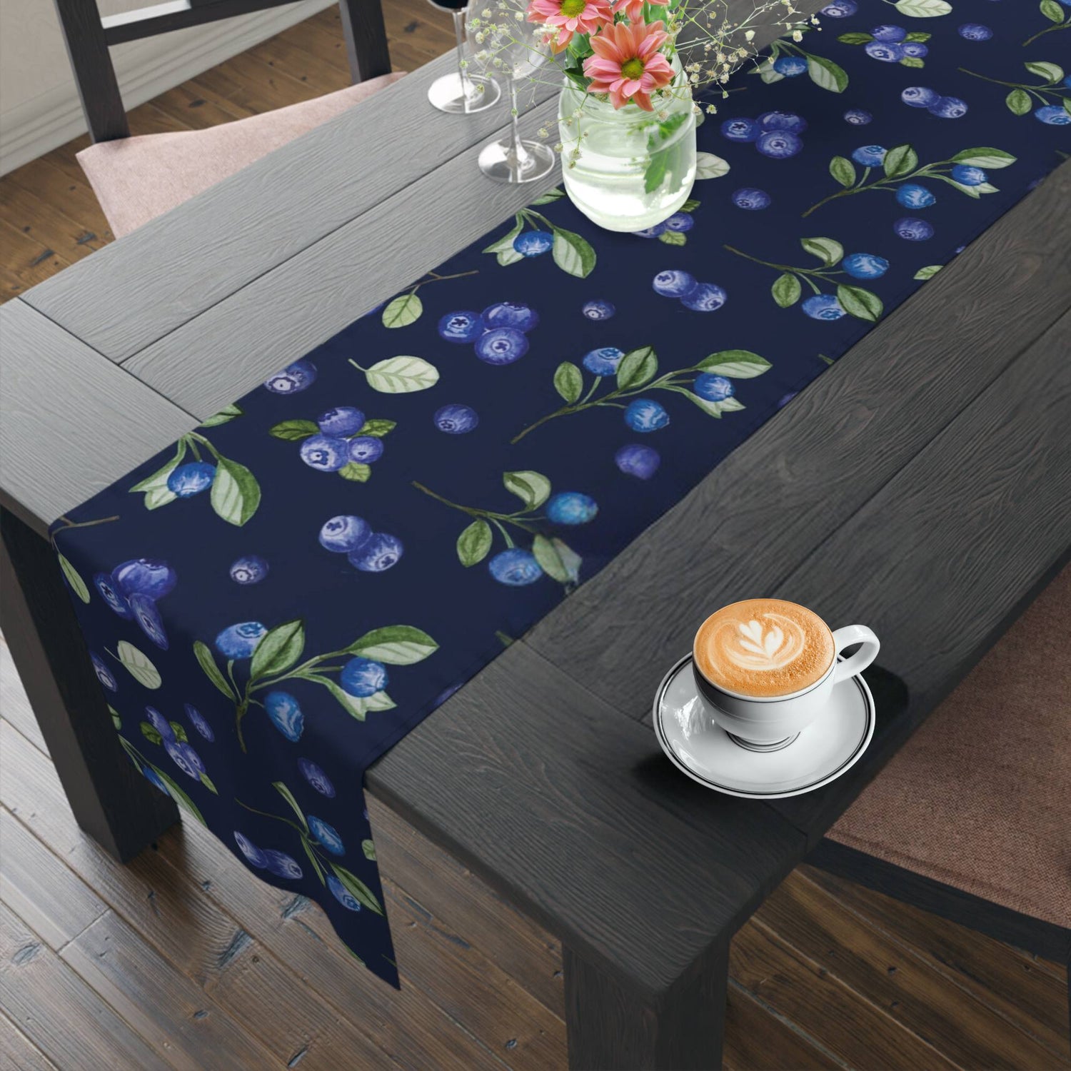 A navy blue table runner with a blueberry print runs along a dark wooden table. A white coffee cup with frothy cappuccino and a vase with pink flowers are on the table.