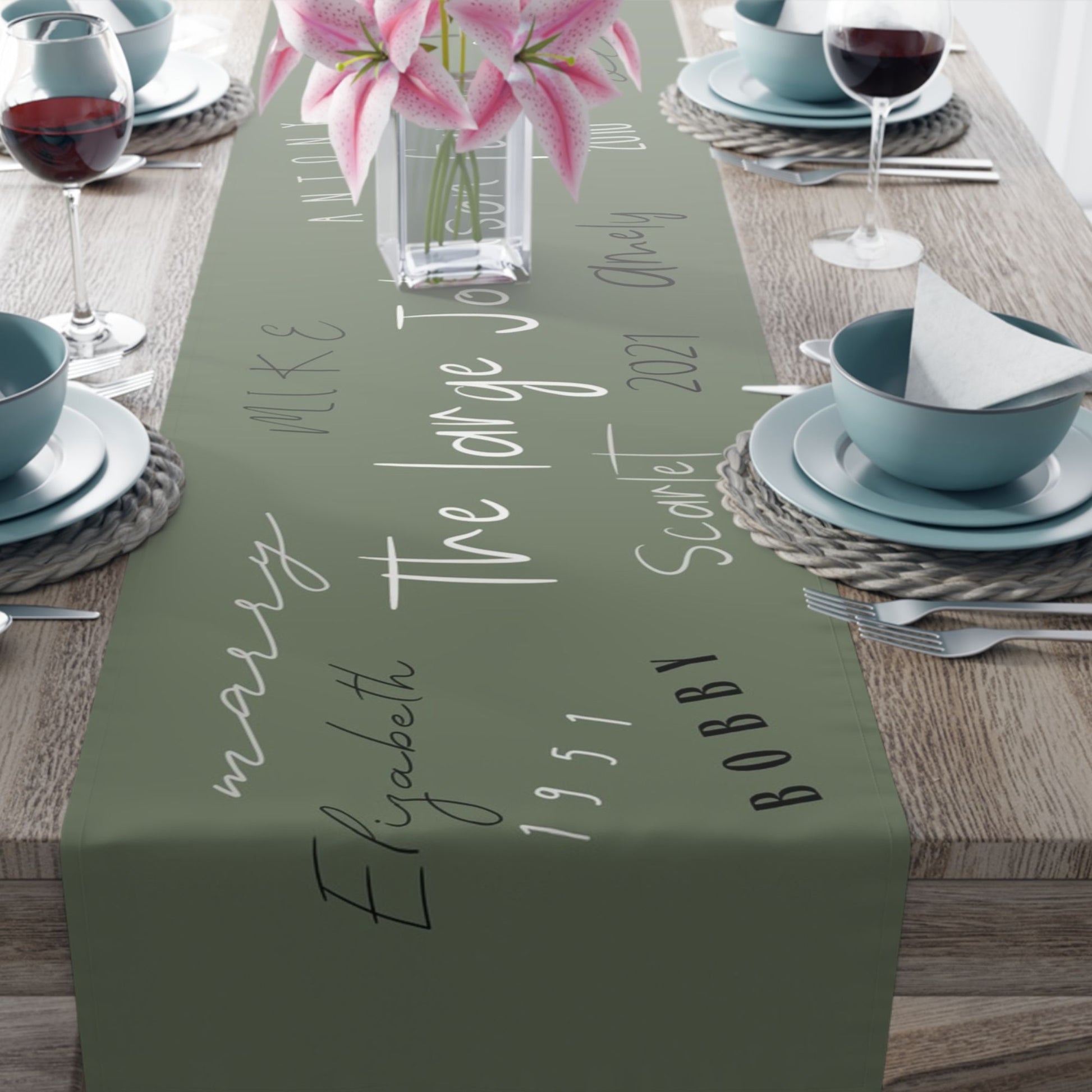 A sage green table runner personalized with various family names written in different styles.