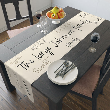 A dark wooden dining table with a cream-colored table runner personalized with various family names written in different styles.