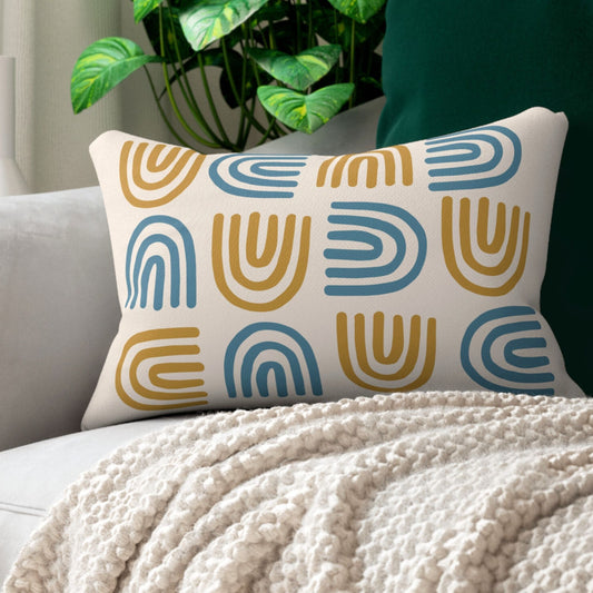Rectangular throw pillow with a modern boho design, featuring alternating mustard yellow and blue arches on a beige background, placed on a light gray couch next to a textured cream blanket.
