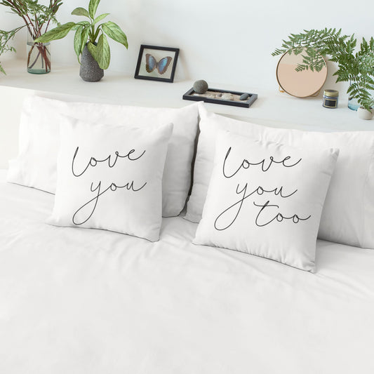 Two cream throw pillows on a bed, one with "love you" and the other with "love you too" written in elegant black script.