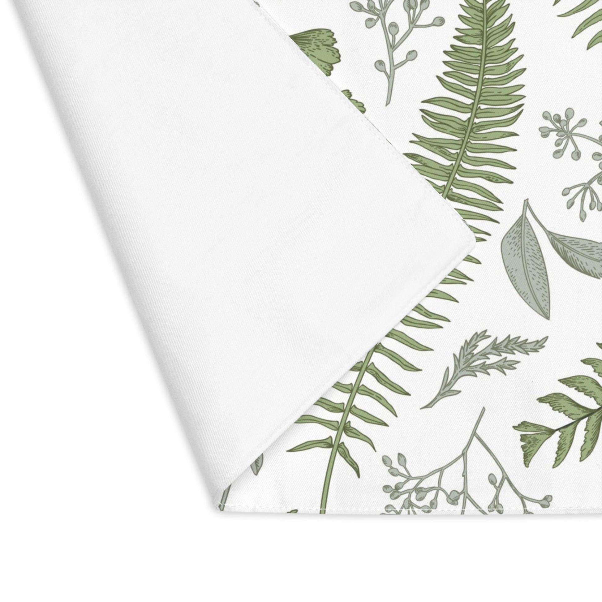 A white placemat with a folded corner, revealing a green botanical pattern featuring various ferns and leaves. The design includes delicate, detailed illustrations of different types of foliage on a white background.